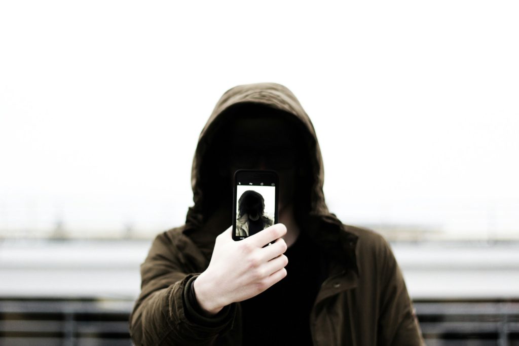 Ein Mann in schwarzer Kapuzenjacke hält ein Smartphone und macht ein Selfie aus der Nähe, mit neutralem Hintergrund und natürlichem Licht.